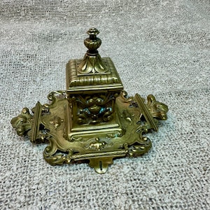 May include: Ornate antique brass inkwell with a square ink pot and decorative flourishes. The inkwell features a detailed design with a small finial on top and an elaborate base with symmetrical patterns. The brass has a warm, aged patina.