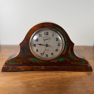 May include: A wooden mantel clock with a rounded top and a silver-toned face. The clock features black Arabic numerals, a smaller sub-dial at the top, and black hands. The wooden frame is decorated with green and gold painted leaf-like accents.