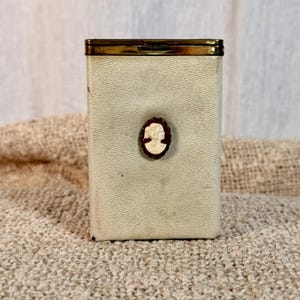 May include: A rectangular off-white textured case with a metallic gold-toned top edge. Centered on the front is an oval-shaped dark brown medallion featuring a raised cream-colored profile cameo of a person facing to the right side.