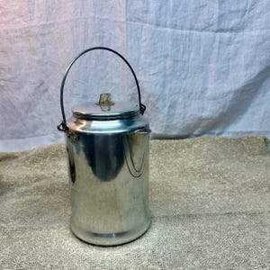 May include: A vintage silver-colored metal camping kettle with a lid and a handle. The kettle has a spout and a small knob on the lid. The handle is a dark metal. The kettle is sitting on a textured surface.