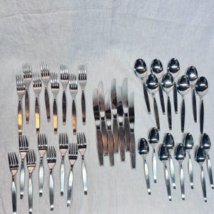 May include: A collection of stainless steel flatware including forks, knives, and spoons arranged in three distinct groups. The set features polished silver-toned metal utensils with simple, straight handles, displayed against a plain white fabric background.