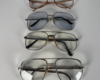 Puede incluir: Cuatro pares de gafas estilo aviador dispuestas verticalmente. El par superior tiene una montura marrón con lentes transparentes. El segundo par presenta una montura plateada con lentes tintadas en azul claro. El tercer y cuarto par tienen monturas plateadas.