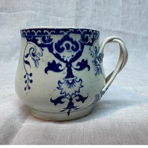 May include: A ceramic mug featuring a white base with a dark blue decorative pattern. The design includes intricate floral and scroll motifs around the rim and body. The mug has a curved handle and a rounded shape, showcasing a classic aesthetic.