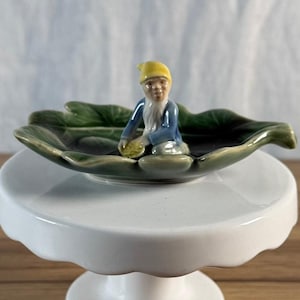May include: A ceramic trinket dish shaped like a green leaf with a small gnome figure sitting in the center. The gnome wears a yellow hat and a blue jacket, holding a small yellow object in its hands. The dish has a glossy finish and detailed veins.