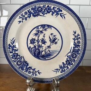 May include: A round ceramic plate featuring a cream-coloured base with intricate blue transferware patterns. The centre displays a circular motif of bamboo, flowers, and a bird, while the rim is decorated with floral vines and geometric borders.