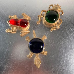 May include: Three gold-toned metal animal brooches. One is a frog with a red cabochon, one is a beetle with a green cabochon, and one is a turtle with a dark blue cabochon. Each piece features a polished metal finish and a central gemstone.