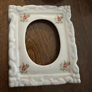 Può includere: Una cornice portafoto rettangolare in ceramica bianca con un bordo sagomato in rilievo e un'apertura centrale ovale. La cornice è decorata con piccoli motivi floreali dipinti di rose rosa con foglie verdi posizionati ai quattro angoli.