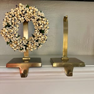 May include: Two brass stocking holders with a metallic finish. One holder features a decorative wreath made of small, round, cream-colored berries with dark centers and green leaves attached to the vertical post of the metal stand.