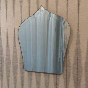 May include: A wall-mounted mirror featuring a distinct onion-dome shape with a pointed top and curved sides. The reflective glass surface has a smooth, frameless edge, creating a clean and simple silhouette that reflects the surrounding environment.