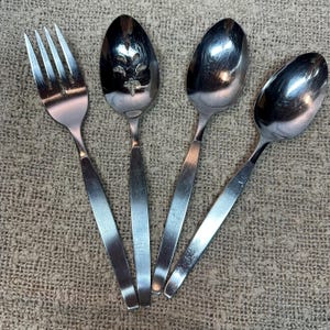 Op de afbeelding: Een set van vier roestvrijstalen bestekstukken, waaronder een vork met vier tanden, een serveerlepel met sleuven en een decoratief uitgesneden patroon, en twee dichte lepels. Elk stuk heeft een gladde, gepolijste afwerking en rechte handgrepen.