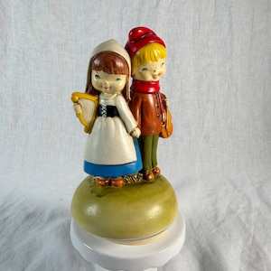 May include: A ceramic figurine featuring two children standing on a rounded green base. One child wears a white dress with a blue skirt and holds a yellow harp. The other child wears a red cap, a brown jacket, and green trousers while holding a violin.