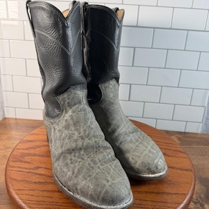 Puede incluir: Un par de botas vaqueras de media caña con cañas de cuero negro con detalles de costuras y empeines de color gris texturizado. Las botas tienen un diseño de punta afilada y tacón, mostrando un esquema de dos tonos con un contraste marcado entre la parte superior negra lisa y la sección inferior gris rugosa.