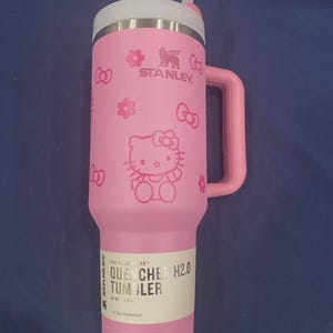May include: Pink Stanley Quencher H2.0 tumbler with a white lid and handle. The tumbler features a Hello Kitty design with bows and flowers. The tumbler has a straw and a label that reads "QUENCHER H2.0 TUMBLER" and "40 OZ".