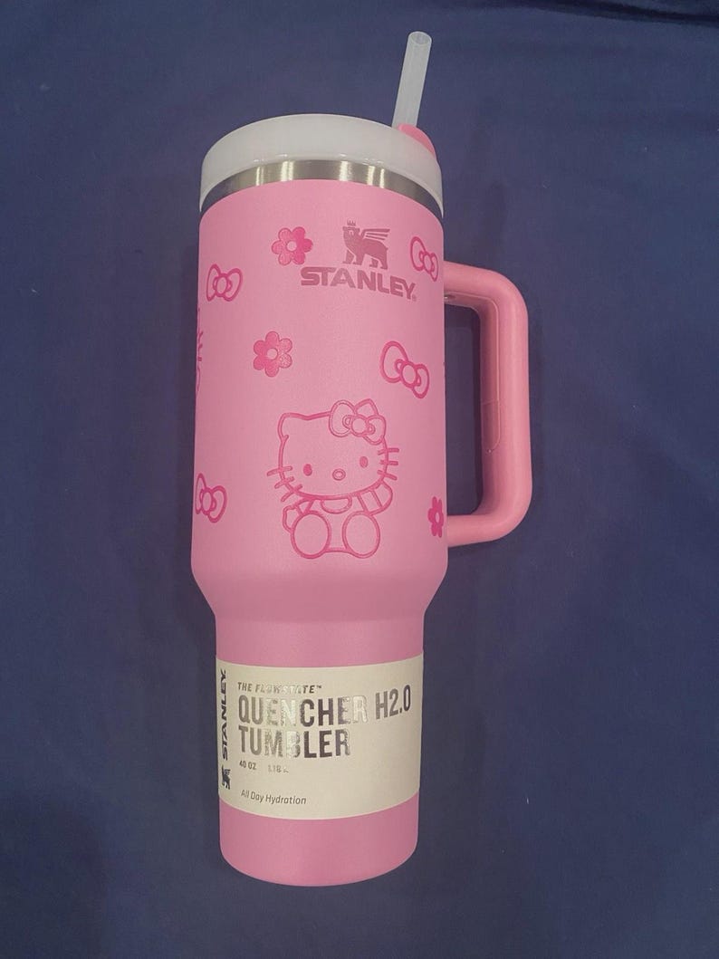 May include: Pink Stanley Quencher H2.0 tumbler with a white lid and handle. The tumbler features a Hello Kitty design with bows and flowers. The tumbler has a straw and a label that reads "QUENCHER H2.0 TUMBLER" and "40 OZ".