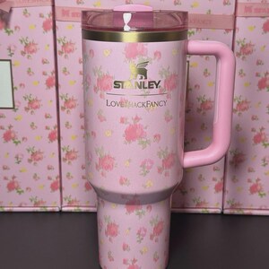 May include: Pink floral Stanley tumbler with a matching handle and lid. The tumbler features the Stanley and LoveShackFancy logos. The background shows boxes with a similar floral pattern.