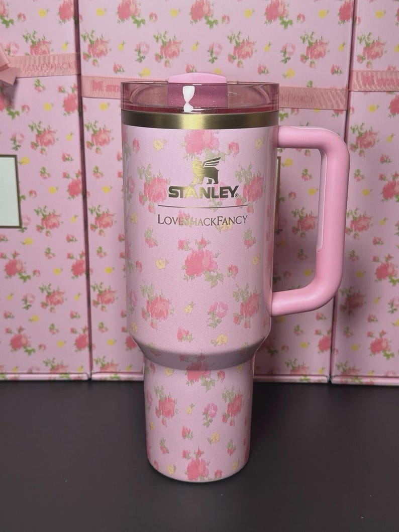 May include: Pink floral Stanley tumbler with a matching handle and lid. The tumbler features the Stanley and LoveShackFancy logos. The background shows boxes with a similar floral pattern.