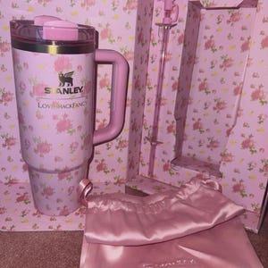 May include: A pink Stanley tumbler with a handle, lid, and floral design, with the text "LOVE SHACK FANCY". The set includes a pink stirring rod, a pink satin bag, and a floral-patterned box.