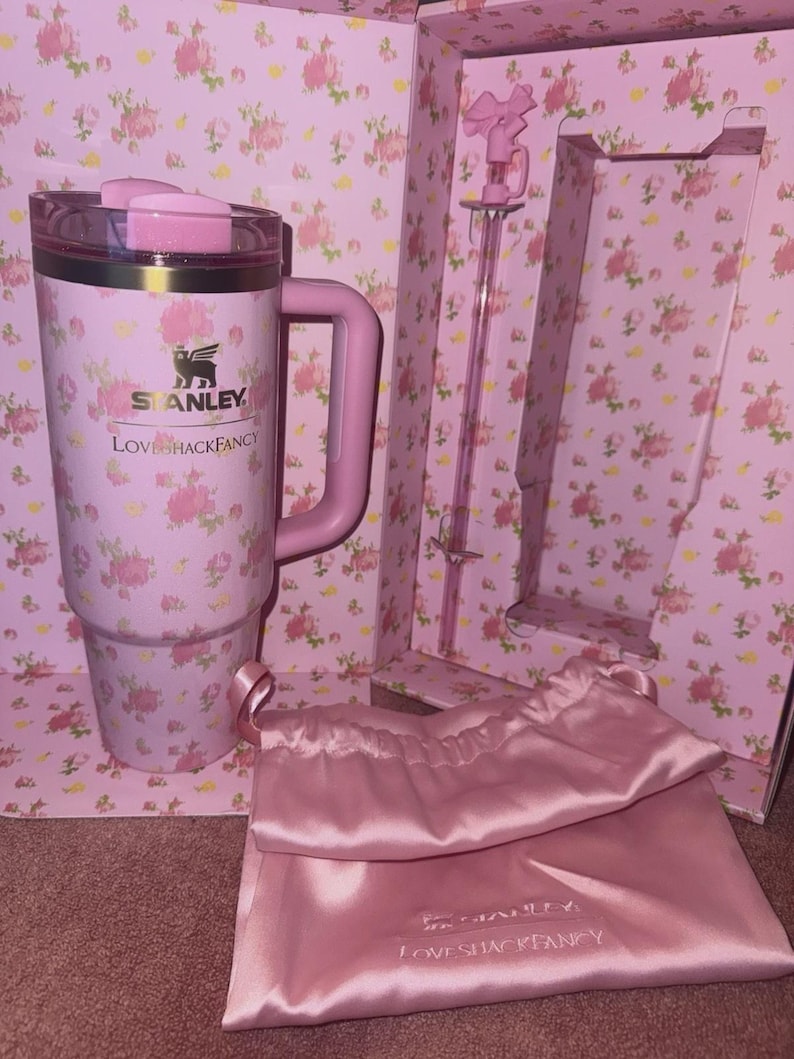 May include: A pink Stanley tumbler with a handle, lid, and floral design, with the text "LOVE SHACK FANCY". The set includes a pink stirring rod, a pink satin bag, and a floral-patterned box.