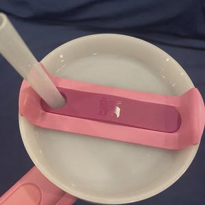 May include: A close-up of a pink and white tumbler lid with a clear straw. The lid has a pink handle and a dark pink accent with a small logo. The straw is inserted into the lid.
