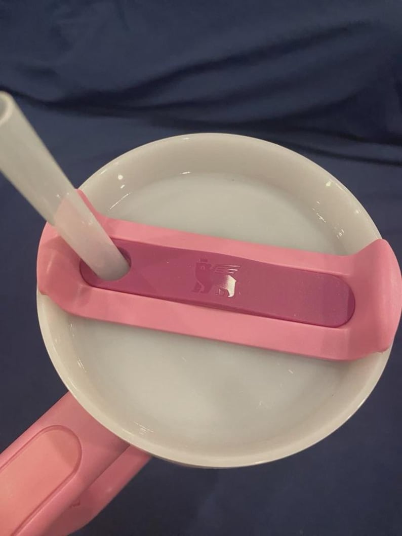 May include: A close-up of a pink and white tumbler lid with a clear straw. The lid has a pink handle and a dark pink accent with a small logo. The straw is inserted into the lid.