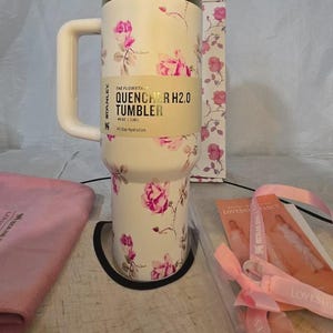 Pu&ograve; includere: Bicchiere Stanley Quencher H2.0 bianco con motivo floreale a rose. Il bicchiere ha un manico e un coperchio. Il testo sul bicchiere recita "QUENCHER H2.0 TUMBLER" e "40 oz | 1,18 L". Viene fornito con un panno rosa e una scatola con un motivo floreale abbinato.