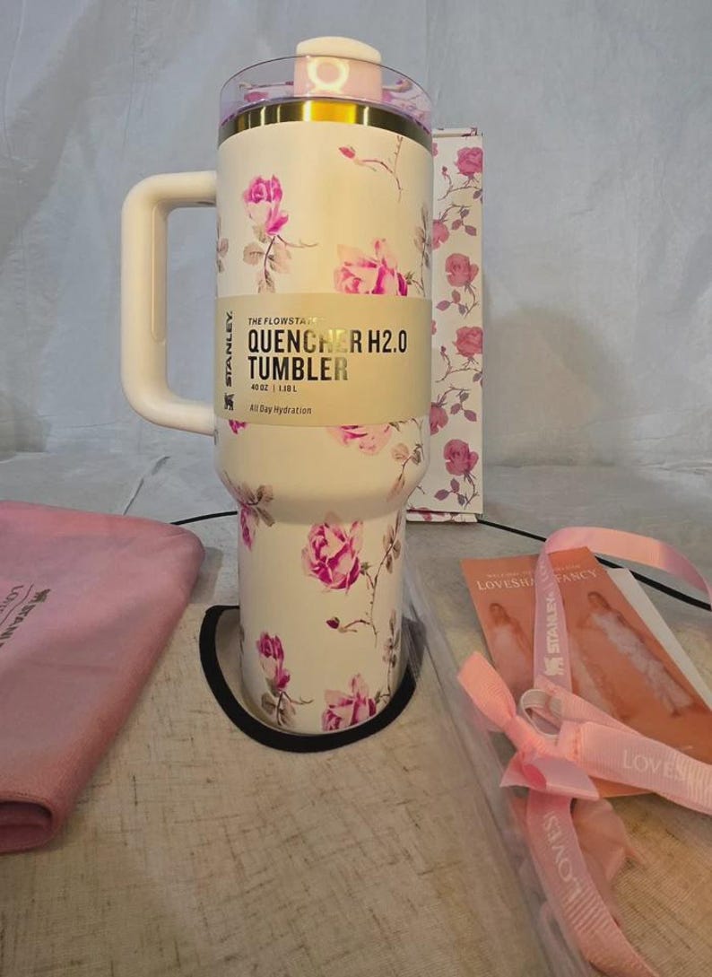 Pu&ograve; includere: Bicchiere Stanley Quencher H2.0 bianco con motivo floreale a rose. Il bicchiere ha un manico e un coperchio. Il testo sul bicchiere recita "QUENCHER H2.0 TUMBLER" e "40 oz | 1,18 L". Viene fornito con un panno rosa e una scatola con un motivo floreale abbinato.