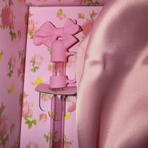 May include: A pink decorative bottle stopper with a bow-shaped handle, set inside a pink floral-patterned box. The stopper has a clear, cylindrical body and a pink base. A pink satin fabric is draped on the right side.