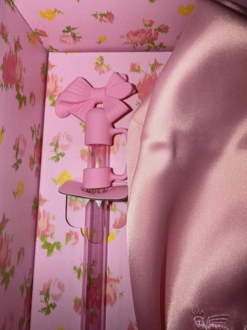 May include: A pink decorative bottle stopper with a bow-shaped handle, set inside a pink floral-patterned box. The stopper has a clear, cylindrical body and a pink base. A pink satin fabric is draped on the right side.