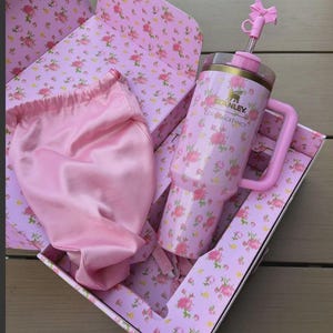 May include: A pink floral Stanley tumbler with a handle and straw, presented in a matching floral box. A pink satin bag is also included. The tumbler features the Stanley logo and the text "Love Shack Fancy".