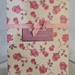 Pu&ograve; includere: Una scatola rettangolare con un motivo floreale di rose rosa su sfondo bianco. Un nastro rosa con un fiocco e un'etichetta con la scritta "LOVE HACKFANCY" avvolge la scatola. &Egrave; una confezione regalo.
