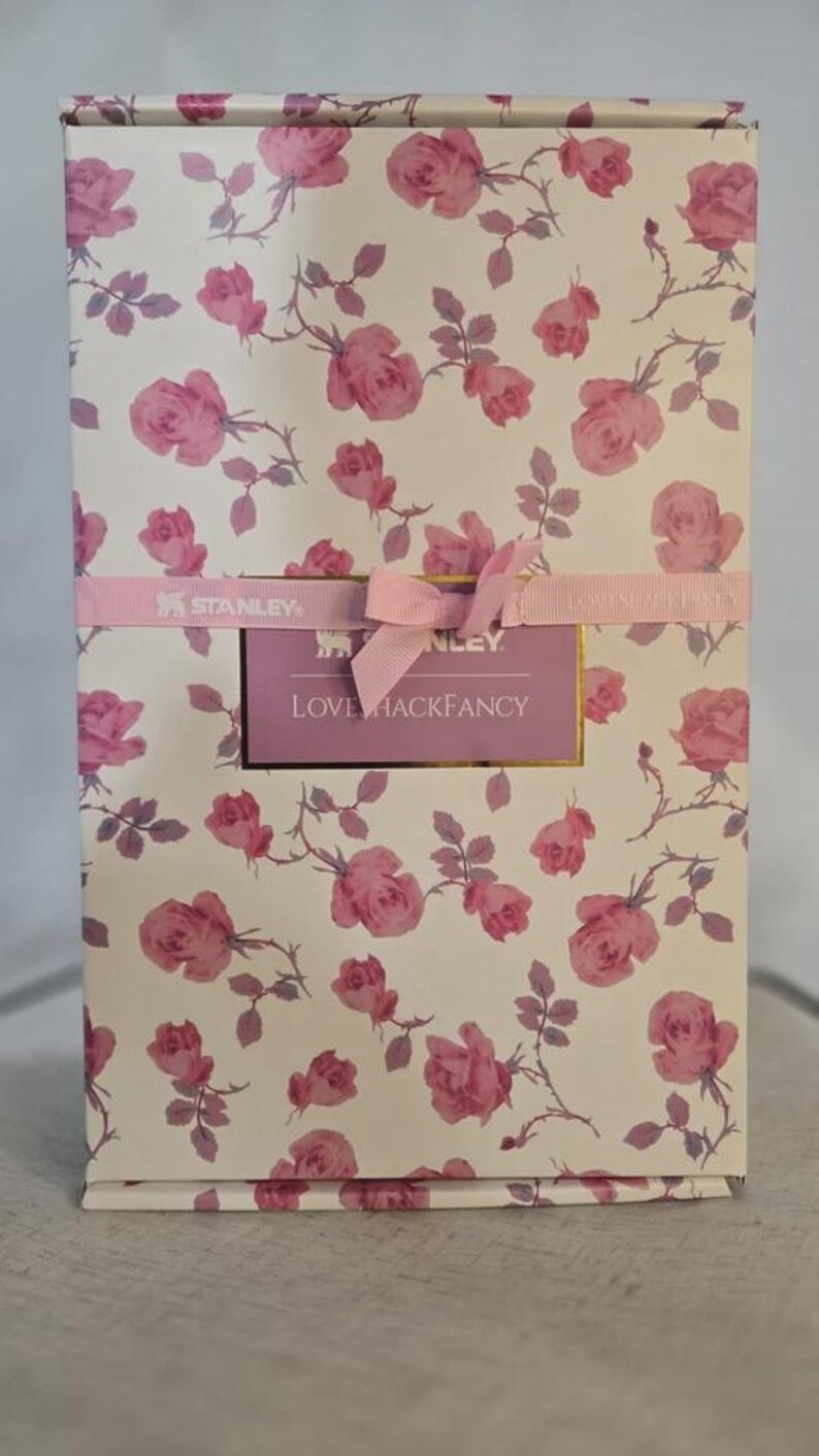 Pu&ograve; includere: Una scatola rettangolare con un motivo floreale di rose rosa su sfondo bianco. Un nastro rosa con un fiocco e un'etichetta con la scritta "LOVE HACKFANCY" avvolge la scatola. &Egrave; una confezione regalo.