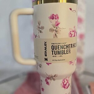 Pu&ograve; includere: Bicchiere Stanley Quencher H2.0 floreale bianco con manico. Il bicchiere presenta un motivo a rose rosa e un bordo dorato. L'etichetta recita "40 oz | 1,18 L".