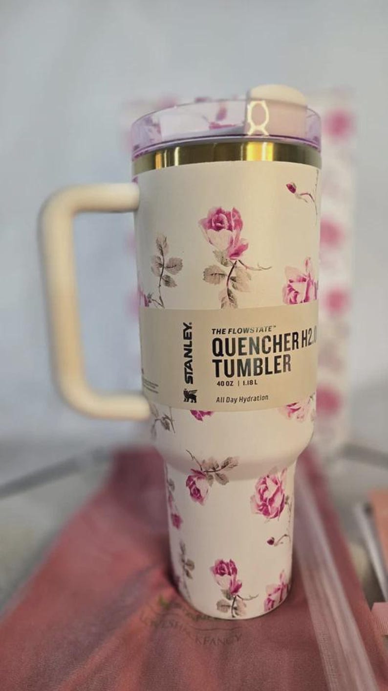 Pu&ograve; includere: Bicchiere Stanley Quencher H2.0 floreale bianco con manico. Il bicchiere presenta un motivo a rose rosa e un bordo dorato. L'etichetta recita "40 oz | 1,18 L".