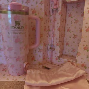 May include: A pink Stanley Loveshackfancy travel mug with a handle, lid, and floral pattern. The mug is in a box with a matching floral design and includes a pink satin bag and a stirring rod.