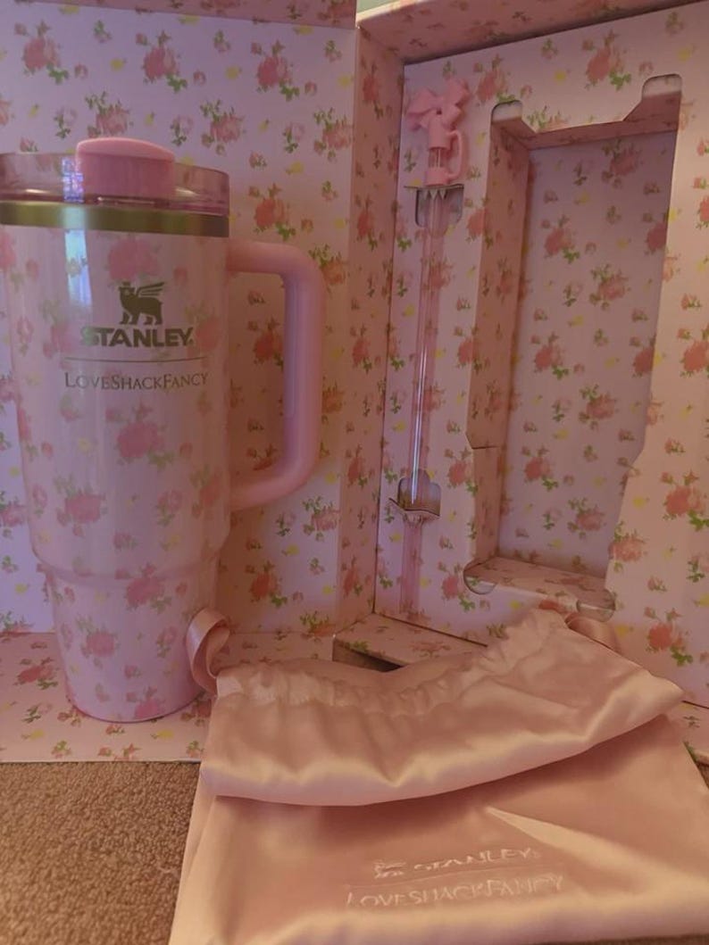 May include: A pink Stanley Loveshackfancy travel mug with a handle, lid, and floral pattern. The mug is in a box with a matching floral design and includes a pink satin bag and a stirring rod.
