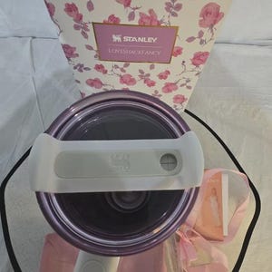Pu&ograve; includere: Un bicchiere Stanley Loveshackfancy viola e bianco con un manico bianco, un sacchetto con coulisse rosa e una confezione regalo con un motivo floreale a rose. La confezione regalo presenta i loghi Stanley e Loveshackfancy.