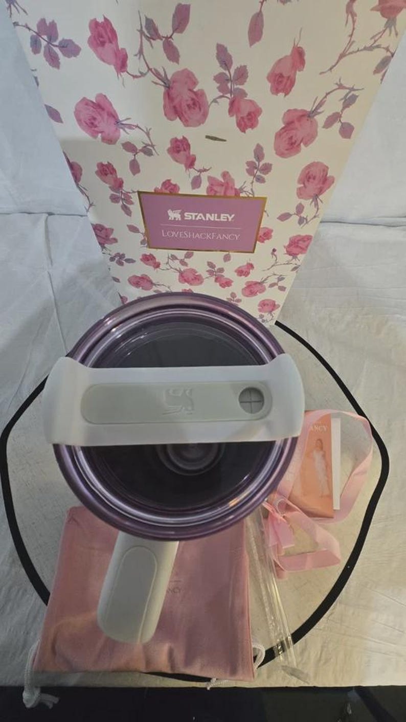Pu&ograve; includere: Un bicchiere Stanley Loveshackfancy viola e bianco con un manico bianco, un sacchetto con coulisse rosa e una confezione regalo con un motivo floreale a rose. La confezione regalo presenta i loghi Stanley e Loveshackfancy.