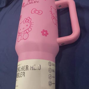 May include: Pink Stanley Quencher H2.0 tumbler with a handle, featuring Hello Kitty graphics and the Stanley logo. The tumbler has a white lid and a pink handle. The text "ENCHER H2.0 TUMBLER" is visible on a white label.
