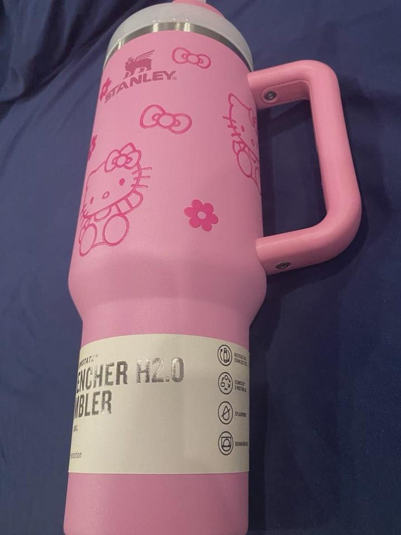 May include: Pink Stanley Quencher H2.0 tumbler with a handle, featuring Hello Kitty graphics and the Stanley logo. The tumbler has a white lid and a pink handle. The text "ENCHER H2.0 TUMBLER" is visible on a white label.