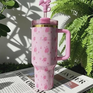 May include: A pink insulated tumbler with a floral pattern and a pink handle. The tumbler has a clear lid and a pink bow-shaped topper. The tumbler is sitting on a newspaper.