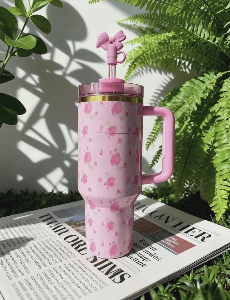 May include: A pink insulated tumbler with a floral pattern and a pink handle. The tumbler has a clear lid and a pink bow-shaped topper. The tumbler is sitting on a newspaper.