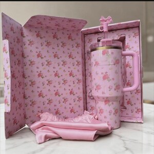May include: A pink floral-patterned tumbler with a handle and straw, presented in a matching gift box. The box and tumbler feature a delicate rose design. A pink satin scarf is also included.