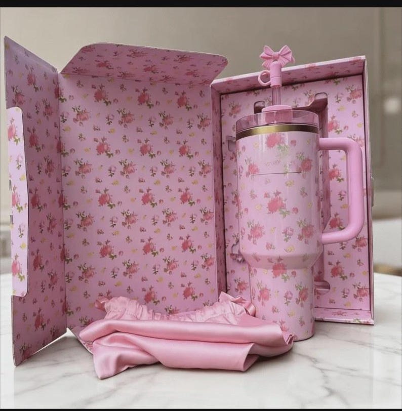 May include: A pink floral-patterned tumbler with a handle and straw, presented in a matching gift box. The box and tumbler feature a delicate rose design. A pink satin scarf is also included.