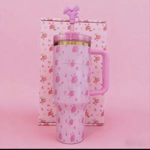May include: A pink floral-patterned tumbler with a handle and a bow-shaped top. The tumbler is paired with a matching box, both adorned with pink roses. The handle and bow are a solid pink color. The tumbler has a gold rim.