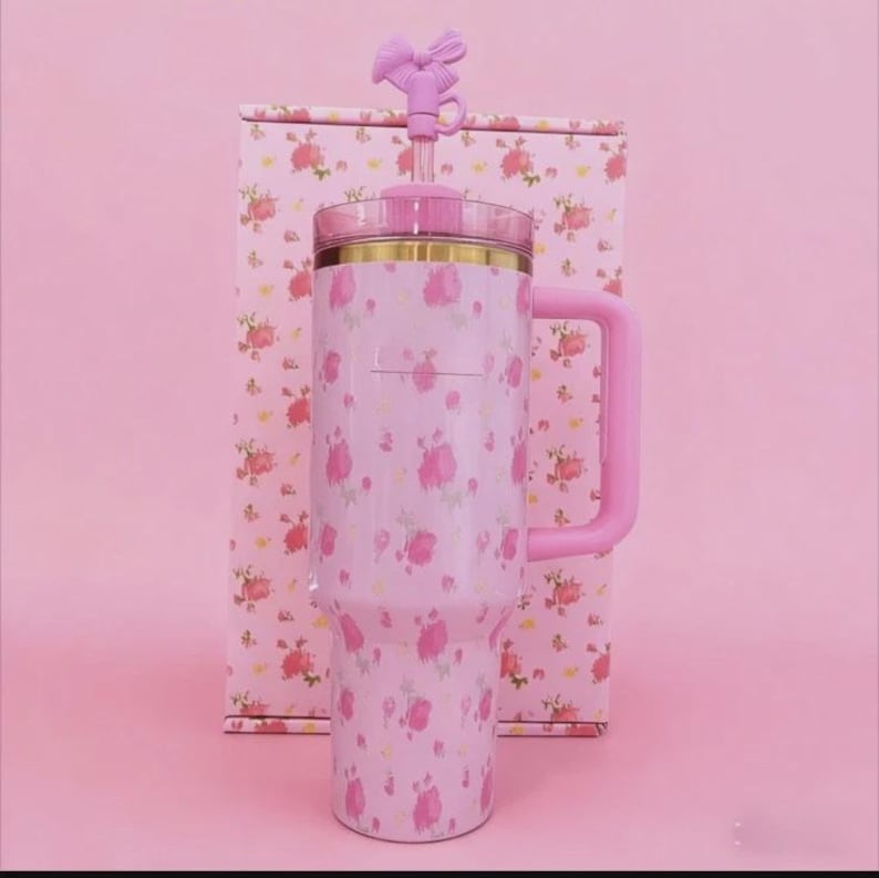 May include: A pink floral-patterned tumbler with a handle and a bow-shaped top. The tumbler is paired with a matching box, both adorned with pink roses. The handle and bow are a solid pink color. The tumbler has a gold rim.