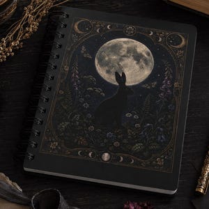 May include: A black spiral-bound notebook featuring a detailed illustration of a rabbit silhouette centered against a large, luminous full moon. The artwork is framed by intricate botanical elements, flowers, and celestial moon phase symbols.