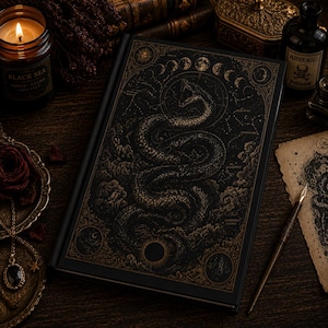May include: A black hardcover book featuring a gold foil illustration of a coiled snake surrounded by celestial motifs, moon phases, and star constellations. The cover design includes intricate patterns and text labels for various astronomical elements.