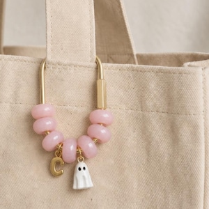 May include: A gold-toned metal carabiner clip attached to a beige canvas tote bag. The clip features six smooth, light pink beads and two small hanging charms: a gold-colored letter C and a white ghost figure with two black eyes.