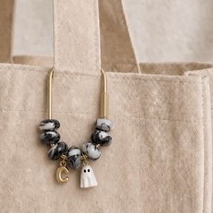 May include: A gold-toned metal carabiner clip attached to a beige canvas tote bag. The clip features a series of black and white marbled beads, a gold-colored letter C charm, and a small white ghost charm with two black eyes hanging from the bottom.