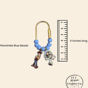 May include: A gold-toned metal keychain featuring a U-shaped clasp adorned with periwinkle blue beads and two character charms. A ruler next to the item indicates it is 4 inches long. The text Periwinkle Blue Beads is visible near the beads.